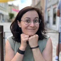 A smiling brown-haired girl with wire-rimmed glasses and an olive sleeveless shirt sits in a open-air café resting her chin on her hands