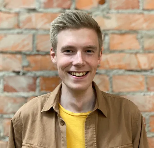 Young man with blond hair wearing a yellow t-shirt and beige shirt is smiling broadly looking at the camera in front of a brick wall