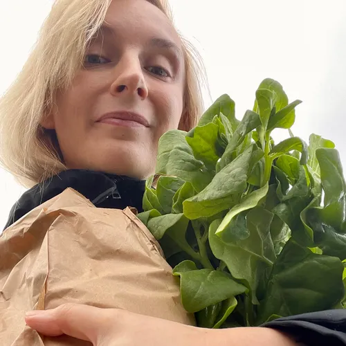 Smiling blond woman in black jacket is looking down at the camera holding a paper bag and bunch of ripe spinach in her arms