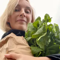 Smiling blond woman in black jacket is looking down at the camera holding a paper bag and bunch of ripe spinach in her arms