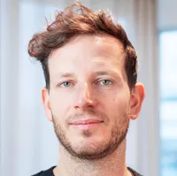 A slightly smiling man with a few days of stubble and short tousled hair looks into the camera in a bright room